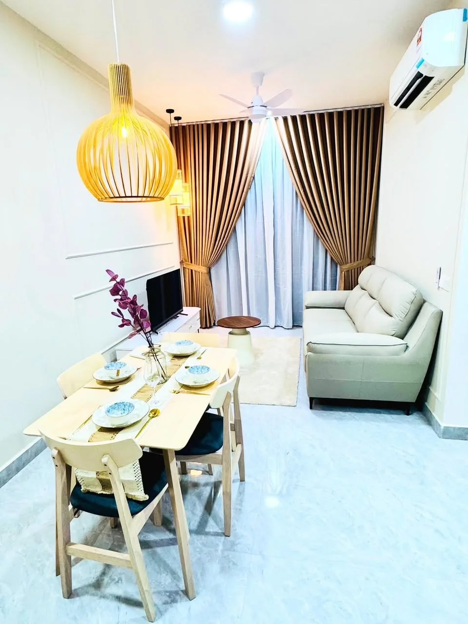 Living features: 3-seater leather sofa in light grey, wooden dining table for 4, pendant lamp with wooden shade, recessed LED downlights, TV console, wall-mounted TV, floor-to-ceiling curtains in beige, light marble flooring, ceiling fan, decorative vase with flowers; style: modern.